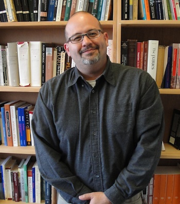 A man in a dark shirt smiles warmly, standing in front of a bookshelf filled with various books.
